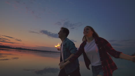 una joven pareja multiétnica feliz sosteniendo velas brillantes y corriendo junto al mar durante la puesta de sol. fotografía en cámara lenta