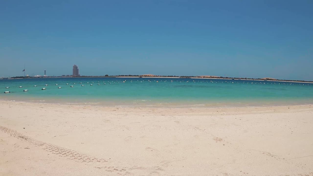 Aerial footage showcases Abu Dhabi's pristine beach with tire-traced sand, buoy-marked waters, and a distant skyline. Captures the serene coastal landscape in vivid clarity and bright lighting