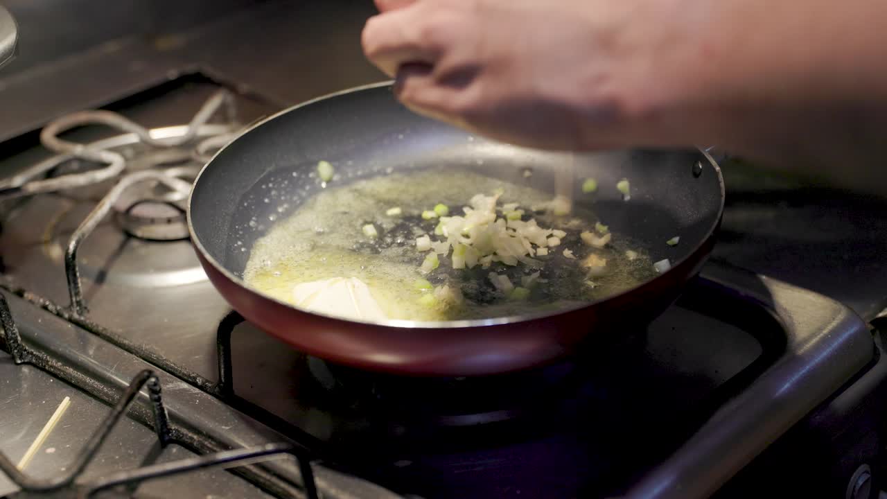 ose-up de una sartén en una estufa con mantequilla derretida chisporroteando suavemente. una mano agrega cebollas picadas en la sartén, creando un chisporroteo satisfactorio a medida que las cebollas comienzan a cocinarse
