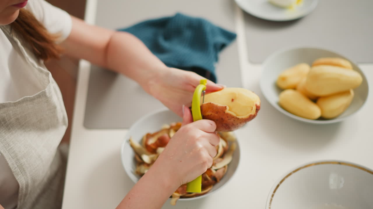 Overhead view of cook peeling potato at white kitchen table, surrounded by peeled potatoes, discarded skins, cloth towel, and plates in clean home kitchen environment
