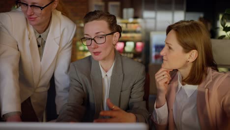 Confident-middle-aged-blonde-girl-with-glasses-in-a-gray-business-uniform-presents-her-work-on-a-laptop-during-a-meeting-of-colleagues-in-a-modern-office