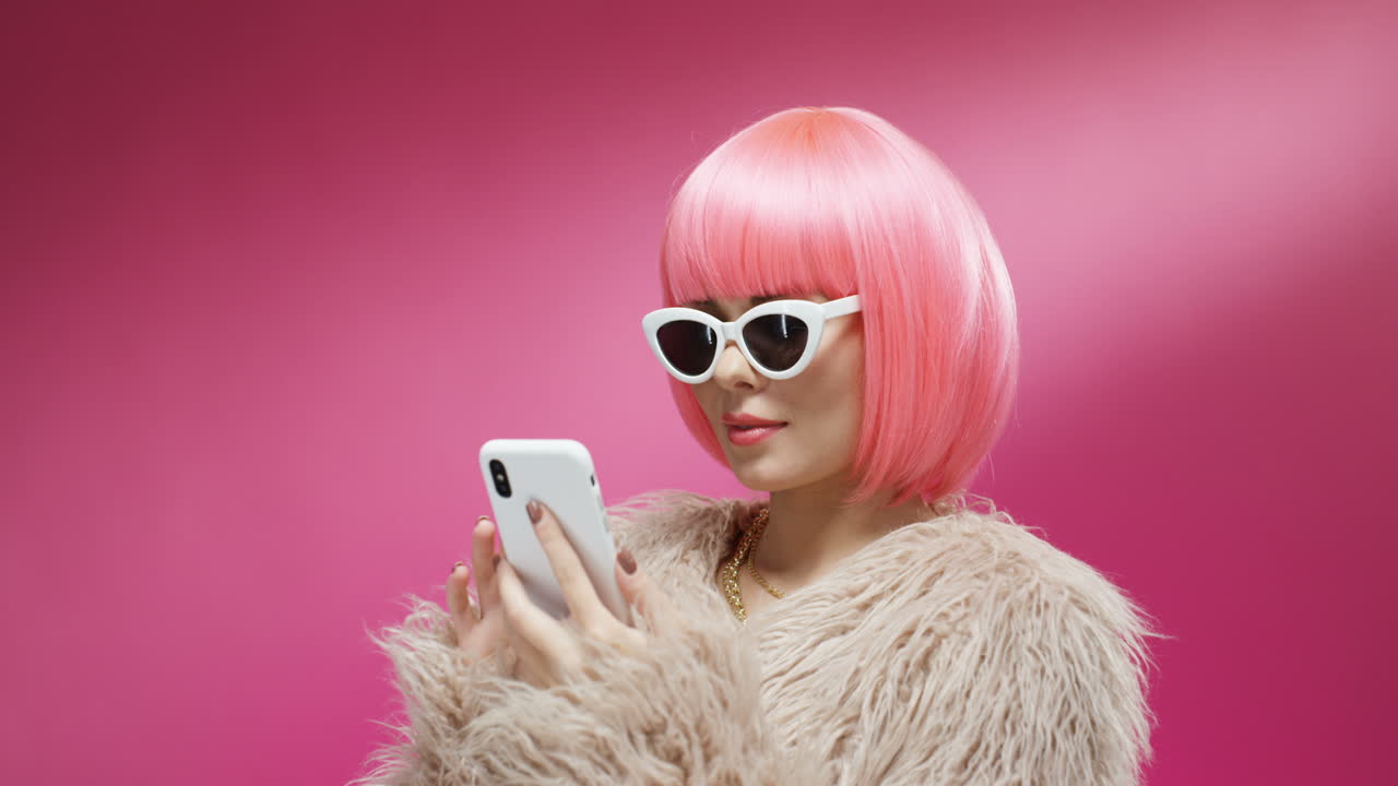 mujer atractiva con gafas de sol y una peluca rosa tocando y enviando mensajes de texto en el teléfono inteligente