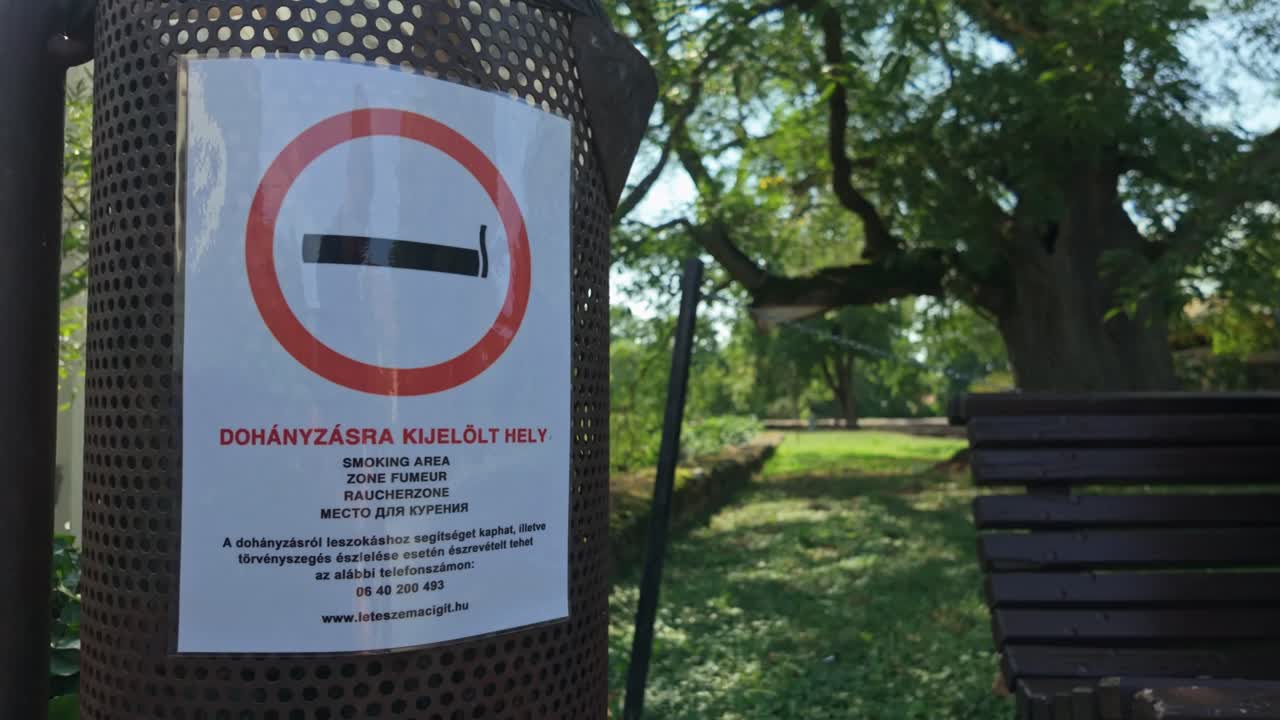 Designated smoking area sign on a metal litter bin inside the park of Castle of Sárospatak, Hungary, surrounded by benches and trees, offering a clear message in a public recreational space