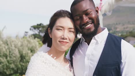 video portrait of happy diverse bride and groom embracing and smiling to camera at outdoor wedding