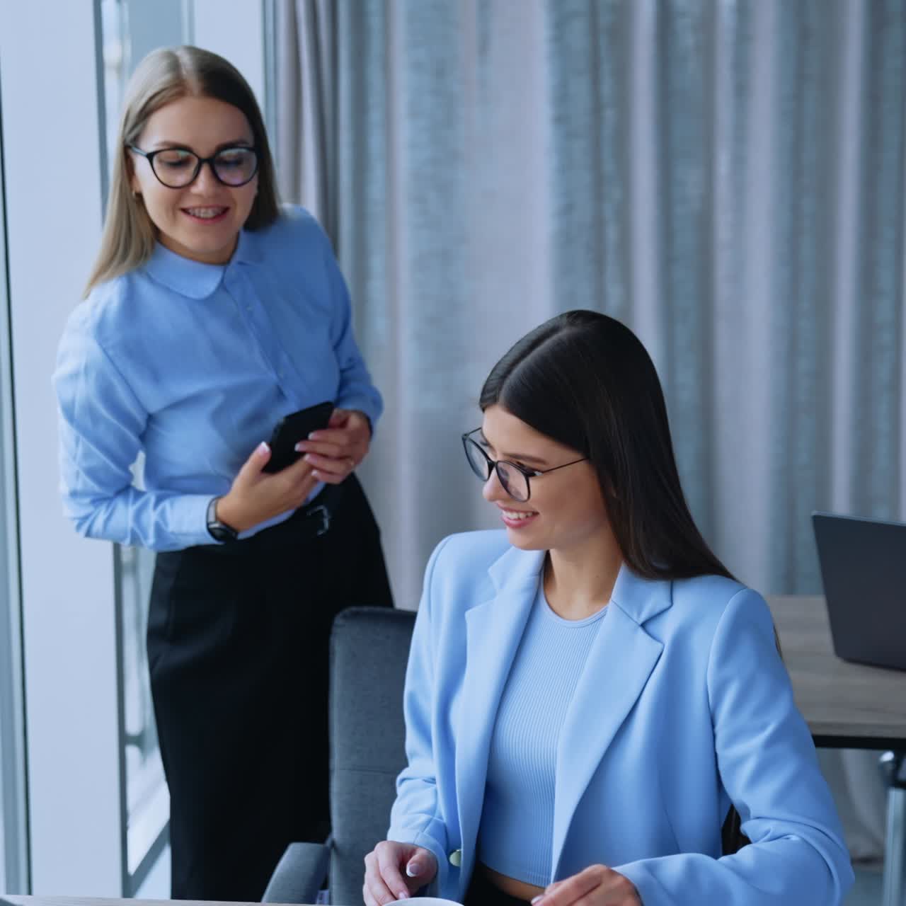 Communication between colleagues at work. Two female employees talking cheerfully and smiling being in the light modern office