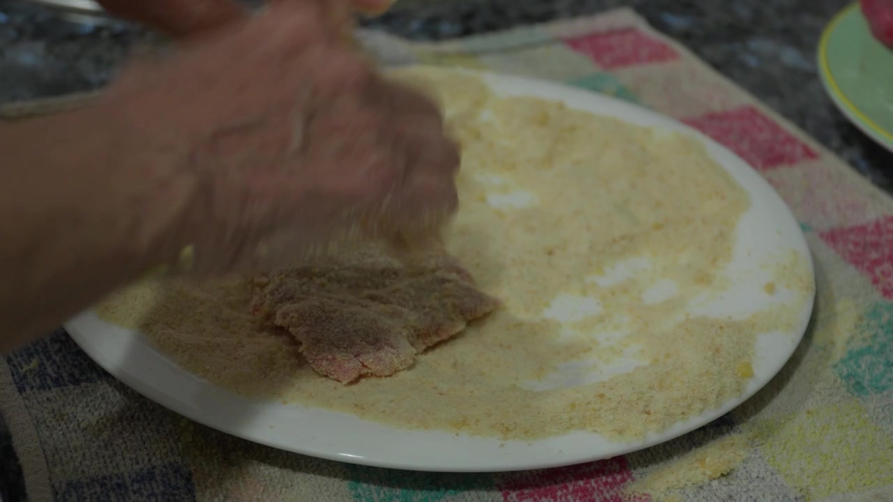 Primo piano delle mani che premono una cotoletta di carne cruda nel pangrattato su un piatto. Preparazione casalinga della classica milanesa argentina
