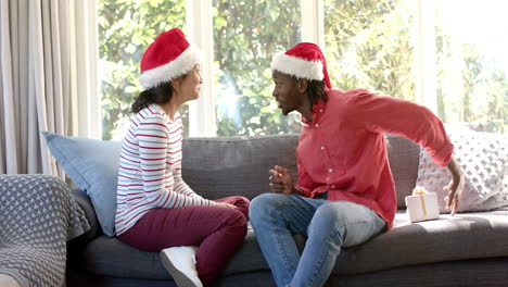 Feliz-Pareja-Diversa-Intercambiando-Regalos-De-Navidad-En-El-Sofá-En-La-Soleada-Sala-De-Estar,-Cámara-Lenta