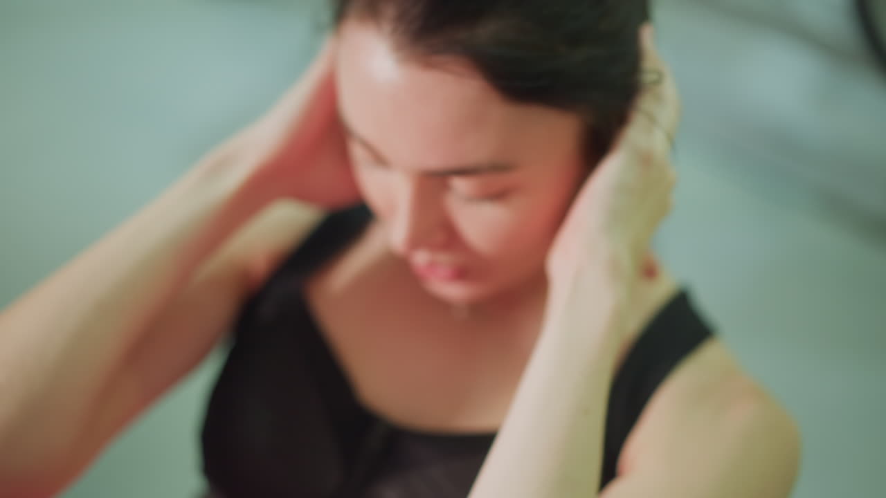 Close-up de mulher recuperando a respiração após uma série, com as mãos nas têmporas, olhos fechados, expressão calma, suor brilhando, inalação e exalação lentas, fundo de estúdio, pausa pós-exercício para regeneração.