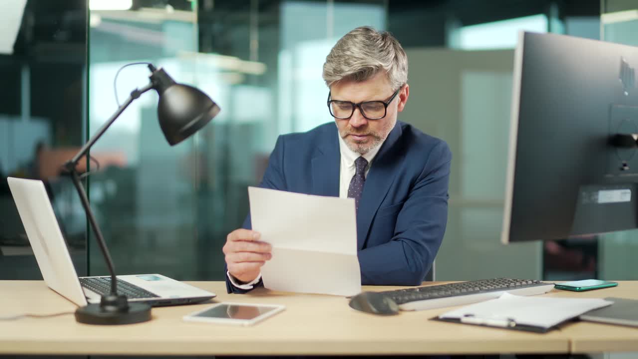 Serious mature businessman in a formal suit examines a document sitting at work in office Gray haired bearded focused business man reads a contract letter or documentation.