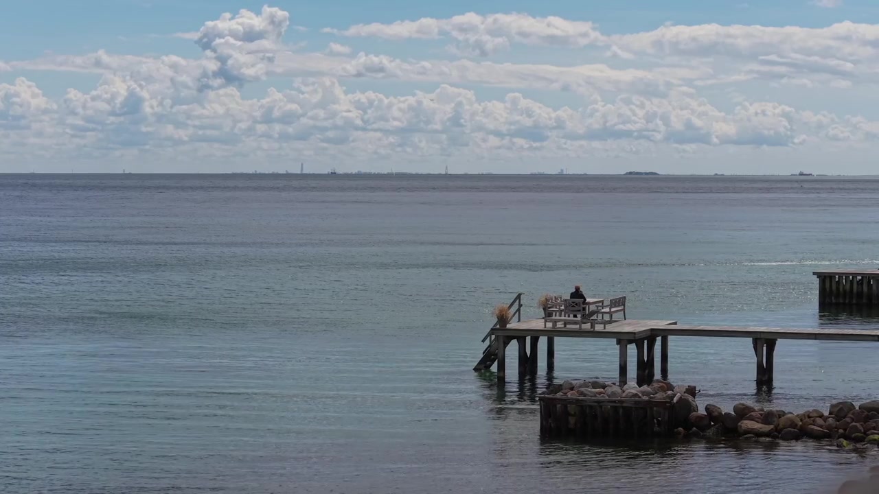 Peaceful footage of an elderly man relaxing on the Taarbaek waterfront promenade, enjoying the calm sea, open horizon, and tranquil coastal atmosphere on Denmark's scenic shoreline.