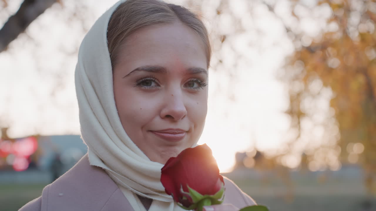 Primo piano di una donna bianca con una rosa. Fiorista che inala il profumo, amante che ricorda, amica che offre un regalo. Controluce del tramonto attraverso rami di betulla, tonalità della pelle delicate, sfondo di un parco cittadino, sorriso tenero, cinematografico.