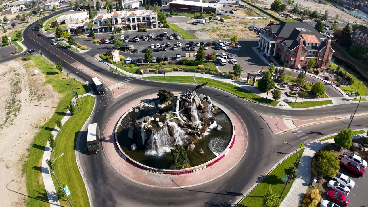 Clip video aerea di un drone che riprende una rotonda circolare a Idaho Falls con strade curve a spirale attorno a un'isola centrale con rocce e vegetazione, circondata da case residenziali e prati curati.