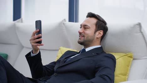 attractive smiling happy bearded businessman uses phone sitting on sofa at home. communication chatting scrolling social media while appearing relaxed in a modern indoor setting