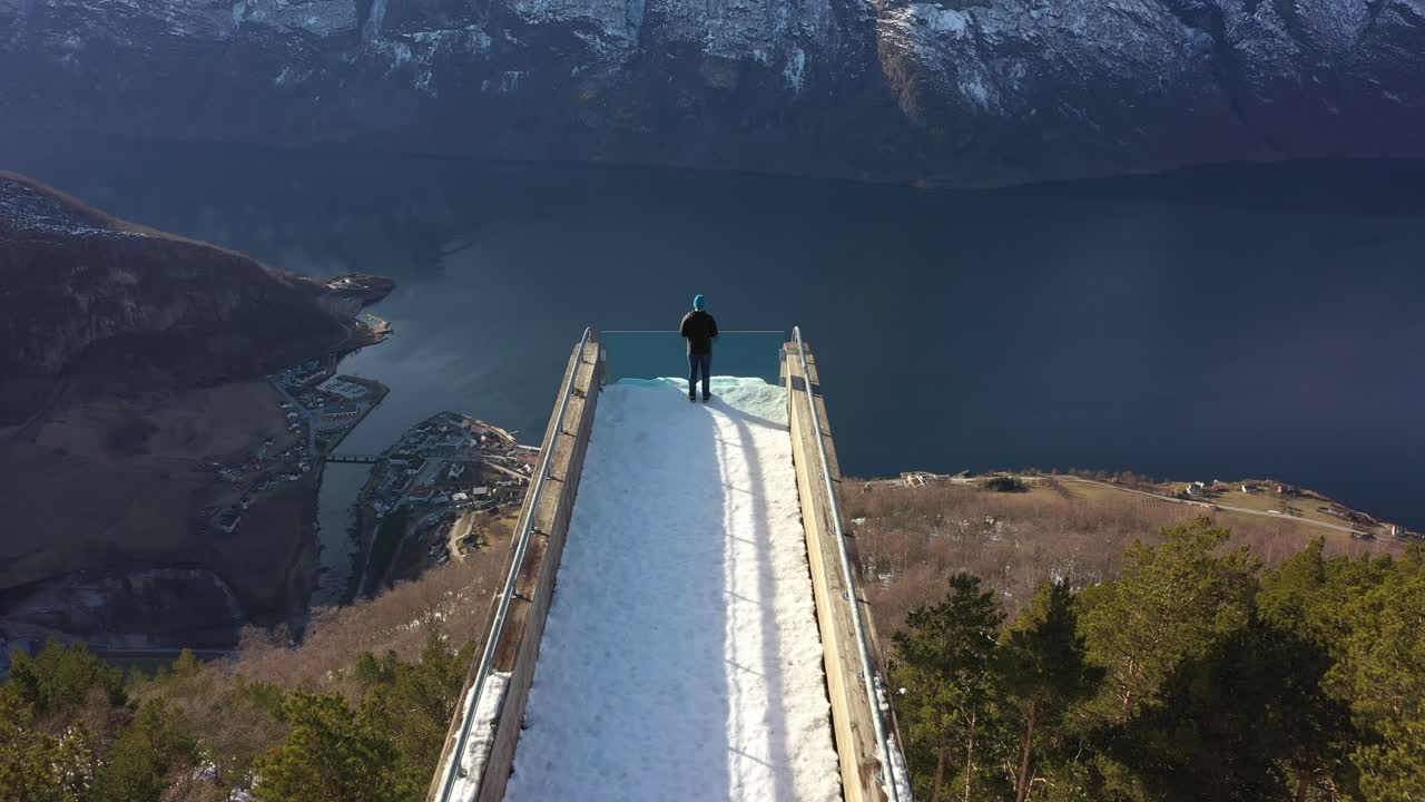 가장자리에 서있는 사람과 함께 스테가스테인 전망대 산책로를 따라 공중 - 천천히 아래로 기울여 언덕 숲과 에울란피요르드 옆에 에울란드 마을을 드러냄 - 화창한 날 노르웨이