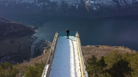 antena a lo largo de la pasarela del mirador de stegastein con un hombre parado en el borde - inclinación lenta hacia abajo para revelar el bosque de la ladera y el pueblo de aurland al lado de aurlandsfjord - día soleado en noruega