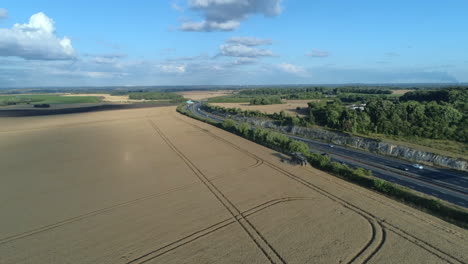 establishing drone shot of combine harvester harvesting on sunny day next to motorway with traffic