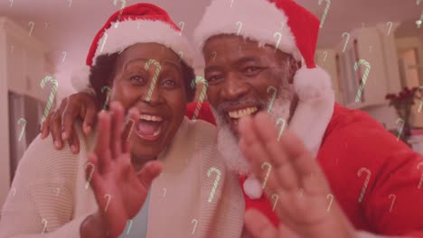 Animation-of-candy-canes-falling-over-smiling-couple-with-santa-hats-waving-hands