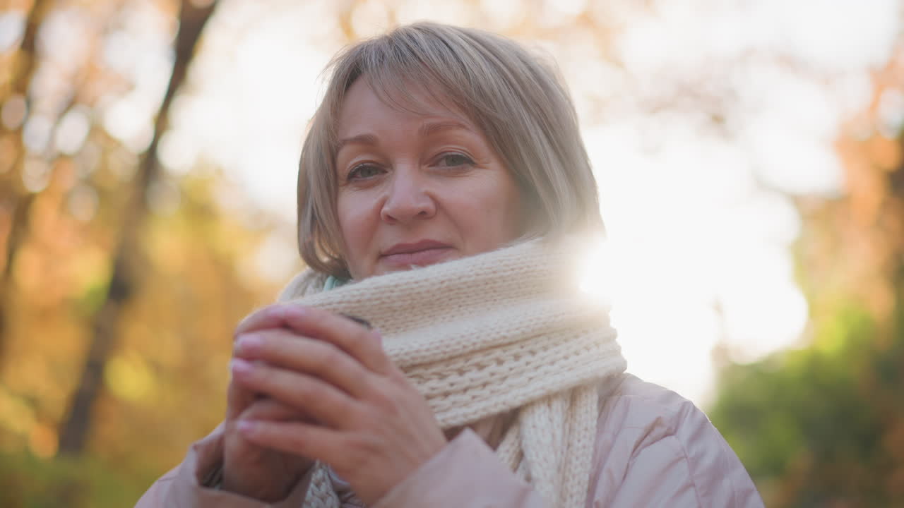elegant mature woman seated outdoors in autumn sunlight, wrapped in chunky knit scarf, cradling cup of tea with both hands as warm glow halo frames head, expression serene and reflective