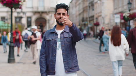 young man smiling friendly at camera, waving hands hello, hi, greeting or goodbye in city street