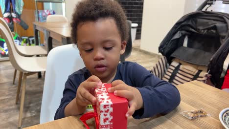 cute two year-old balck baby tries to open a tiny red gift box seated next to his mother inside a cafeteria