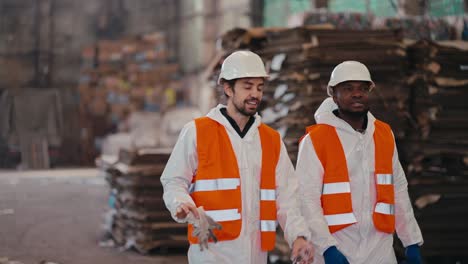 a happy brunette man with a beard walks with his colleague, a man with black skin color and in a white protective uniform in an orange helmet through a large plant with waste paper and plastic for recycling and sorting