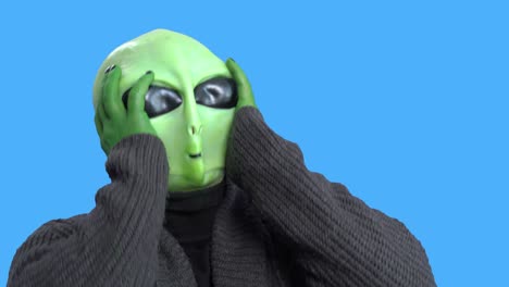 man in alien costume, warm knitted jacket, grabs his head with his hands, writhes with laughter, against a blue chroma key. preparation for halloween party.