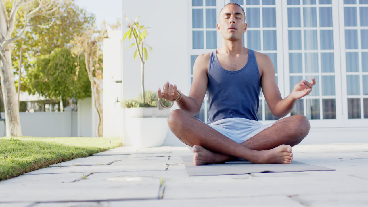 concentrado hombre biracial practicando meditación de yoga en el jardín soleado, cámara lenta