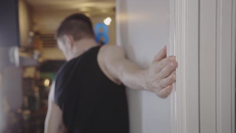 blurry view of handsome caucasian guy inside the house standing, facing the wall and stretching his right arm