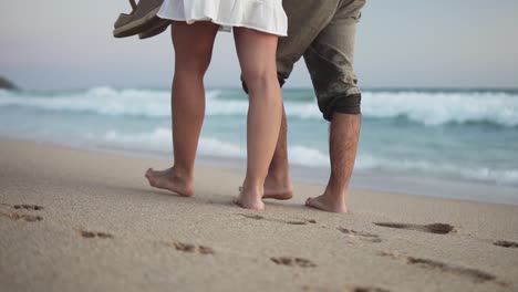 slow motion handheld shot of a couple in love walking along the sand on the beach in front of the sea with calm waves with their dog leaving footprints in the sand