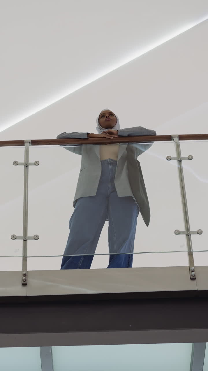 Muslim African-American woman with hijab leans on bridge glass railing between shopping center levels low angle shot. Lady spends time in corporate building