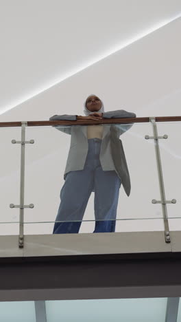 muslim african-american woman with hijab leans on bridge glass railing between shopping center levels low angle shot. lady spends time in corporate building