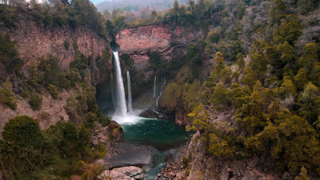 Forward drone aerial of majestic waterfall into emerald pool, Siete Tazas National Park, Ñuble, Chile with red cliffs and autumn forest