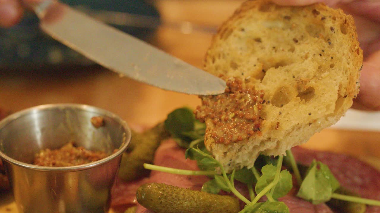 A hand spreads pâté onto a slice of rustic bread beside a charcuterie platter with salami, cheese, pickles, and fresh greens under warm lighting