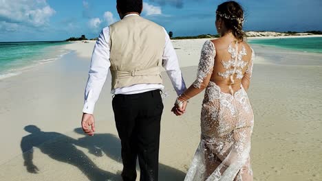 Recién-Casados-Caminando-Vistos-Desde-Atrás-En-La-Playa-De-Arena-Blanca,-Isla-Caribeña-Con-Salpicaduras-De-Agua-De-Mar