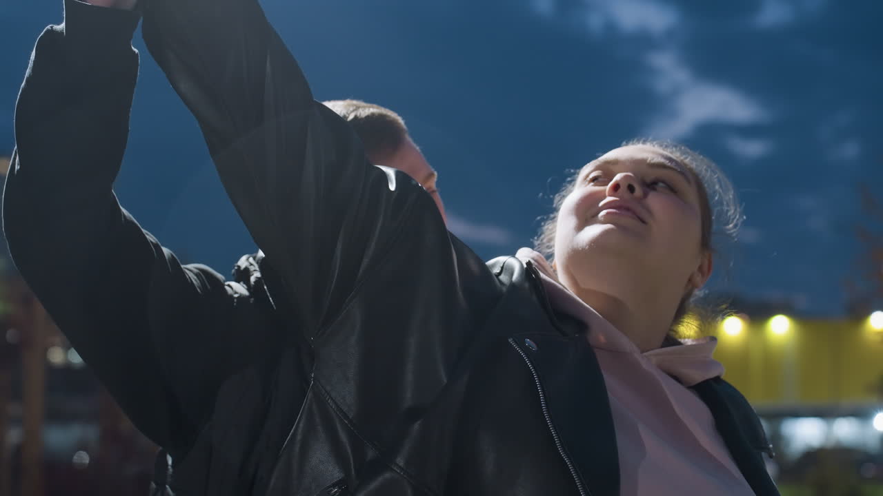 Close-up of woman looking at her man as they dance with light reflecting off their faces, residential buildings softly illuminated in background under night sky