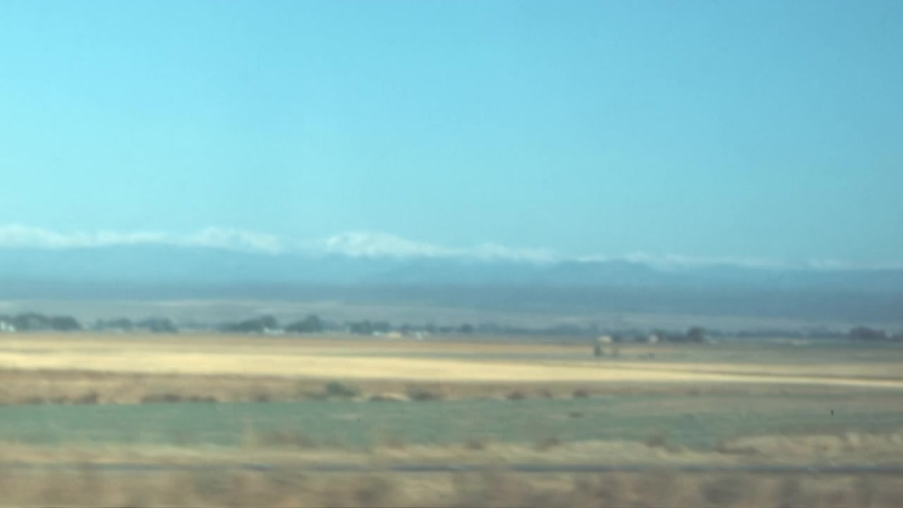 Un veicolo percorre un'ampia valle arida vicino a Los Angeles, Stati Uniti, negli anni '60. Macchia mediterranea e campi costeggiano un'autostrada che conduce a montagne lontane, filmati d'archivio. Rallentatore