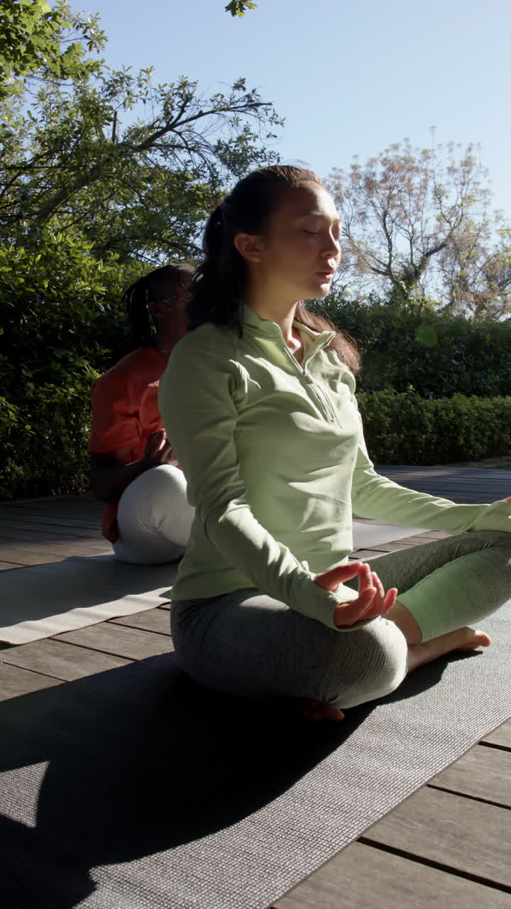Vertical video of diverse couple practicing yoga meditation sitting out in sun, slow motion
