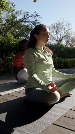 vertical video of diverse couple practicing yoga meditation sitting out in sun, slow motion