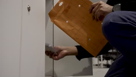 un individuo abre un pequeño armario personal en un entorno de oficina interior, colocando un sobre de correo interno dentro