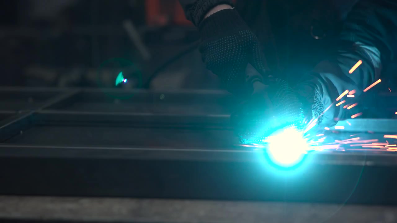 Close view of industrial repair work shows intense sparks and blue white flare at metal contact, with dark blurred background, shallow focus, heavy contrast, and partial worker in safety clothing