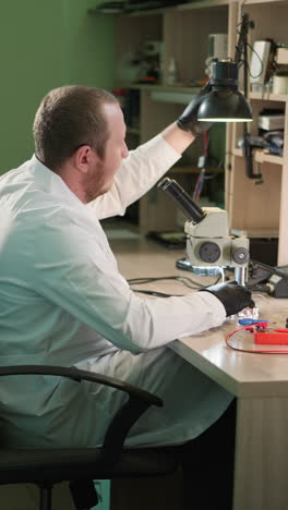 a close view of a technician in a white lab coat and black hand gloves adjusting a desk lamp and continues work circuit under a microscope using a small iron picker with a shelf by the side