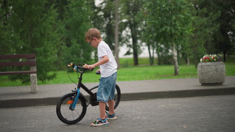 a young boy wearing a white t-shirt and denim shorts prepares to move and climb his black bicycle in a park as he rides off