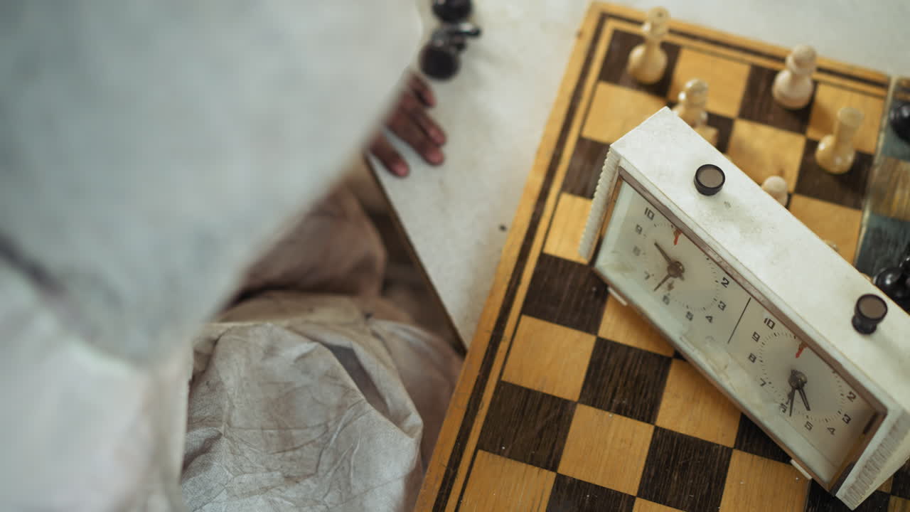 Rear view of kid in white protective clothing playing with chess clock beside chessboard, dirty hands pressing timer during game, showing focus, childhood concentration and atmosphere of serious play