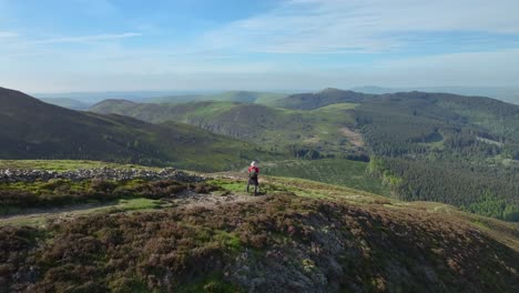 Mountain-walker-on-high-path-with-flyover-revealing-green-valley-and-pine-forested-mountains-to-distant-lowlands-prior-to-sunset