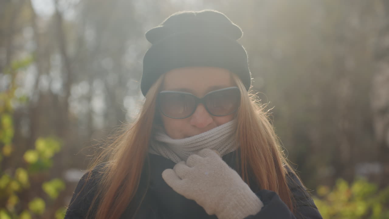 Primo piano di una donna bianca dai capelli rossi che sorride alla luce del sole, indossando occhiali da sole, sciarpa di lana e guanti, con un'espressione giocosa e un gesto della mano malizioso. Uno zaino è visibile sullo sfondo, con una calda retroilluminazione dorata e polvere.