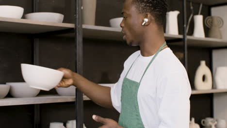 close up of an male clerk with wireless earbuds stocking ceramic bowl on the shelf in the pottery store