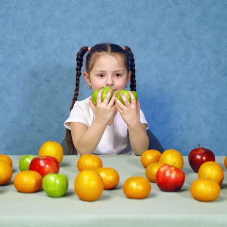 smiling little girl holds two green apples at home. cheerful kid sitting at the table full of fresh organic fruits and playing with apples on blue background.