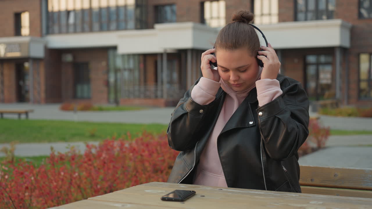 White woman seated outdoors puts headset on and reaches for phone on table with red flowers swaying in wind and modern office building in background during calm afternoon moment