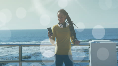 Animation-Von-Lichtpunkten-über-Einer-Frau-Mit-Gemischter-Abstammung,-Die-Auf-Der-Promenade-Ein-Smartphone-Benutzt