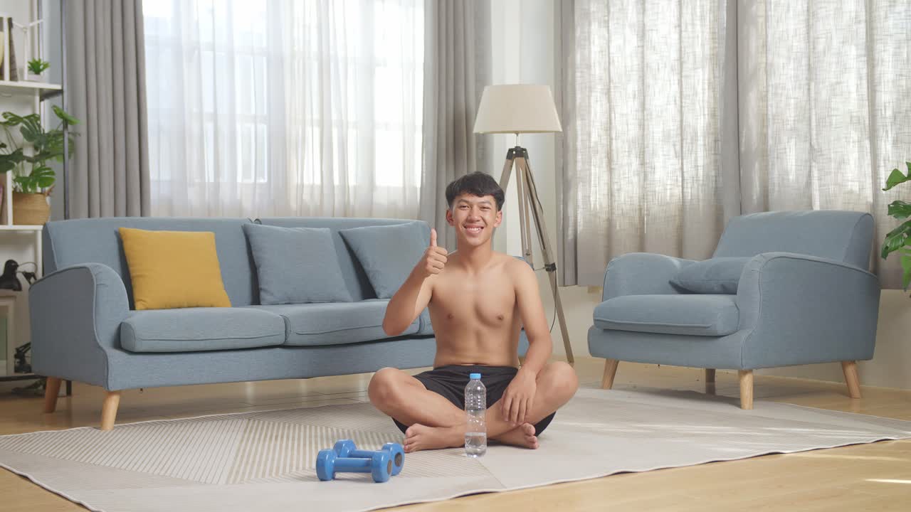집에서 셔츠를 벗고 운동하는 동안 매트에 앉아있는 동안 웃고 엄지손가락을 보여주는 덤벨을 가진 아시아 십대 소년의 몸이 가득 차 있습니다.